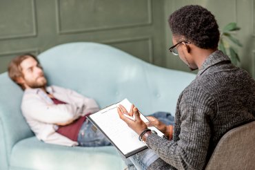 A therapist speaks to a patient with a clipboard in his hand. The male patient lays on a couch with his arms folded, staring at the wall.