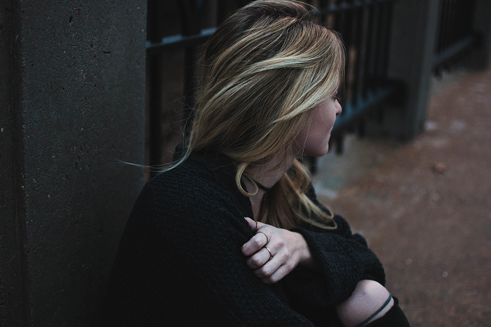 A young woman sits against a fence on the sidewalk, holding herself and looking into the distance.