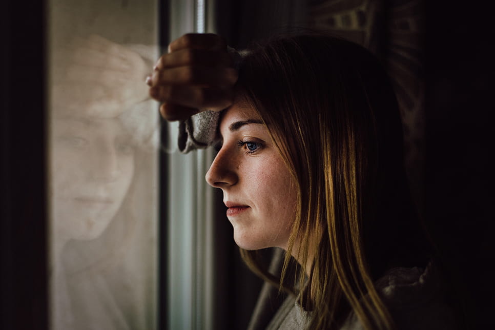 Young woman resting head against the back of her hand, leaning against a window and looking out it.