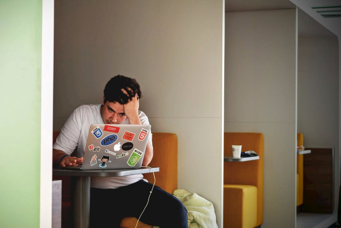 A young man sits at a study table, running his fingers through his hair while he looks at his laptop in stress.
