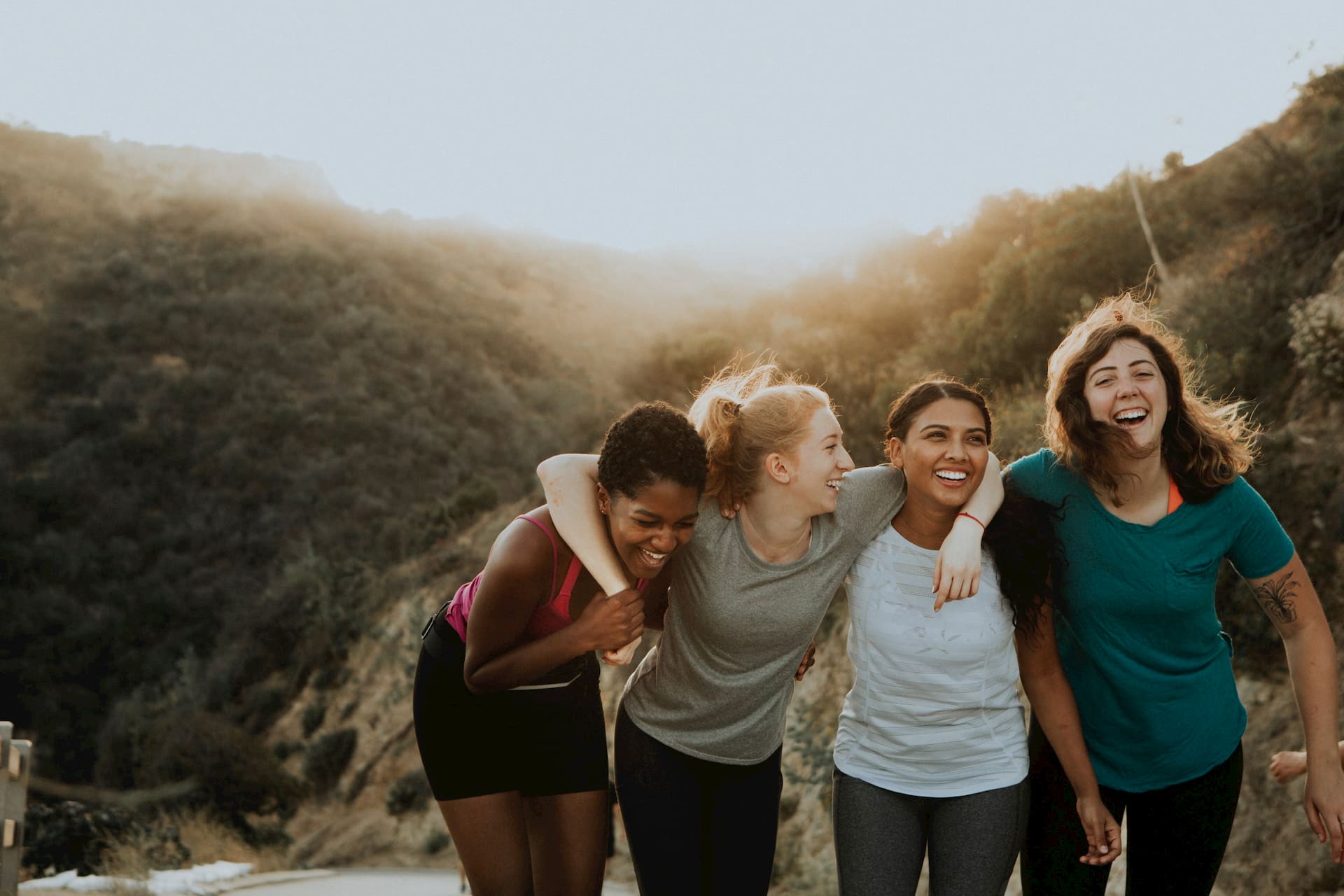 A group of four happy young women embrace in a line out in a hilly landscape at sunrise.