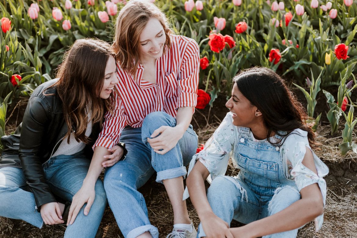 Three young women sit among flowers. They're laughing and enjoying each other's company.